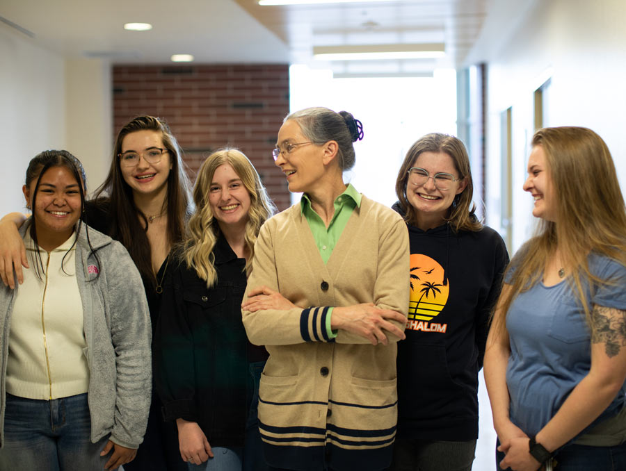 Six women stand at the end of a hallway smiling. A woman in the middle wearing a cardigan is looking to her right and smiling.