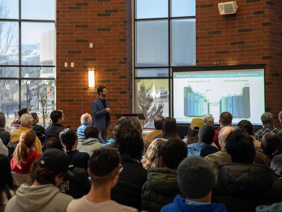 A man stands in front of a crowded room pointing to a screen with two bar graphs on it.