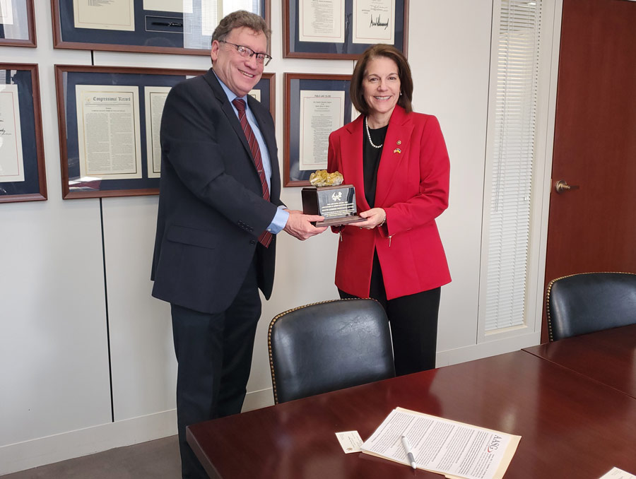 Jim Faulds hands Senator Catherine Cortez Masto an award. The award is a block of wood with a plaque and a piece of a gold-colored rock on it. They are both dressed formally and are smiling for the photo, being taken in an office.