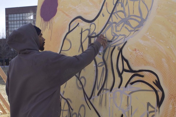 Artist Doomed Movement, Terrence Hammond, works with a spray can in hand on a large yellow mural outside.