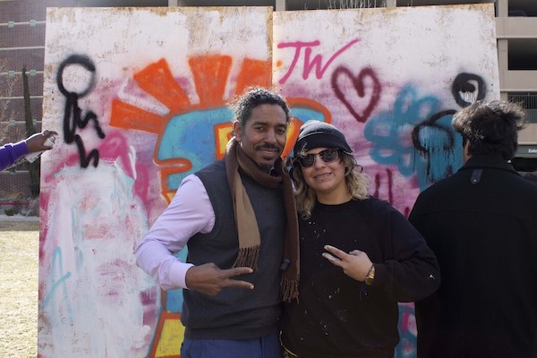 Dr. Frederick Gooding and a student graffiti artist pose in front of a collaborative mural, holding up sideways peace signs with their fingers. 