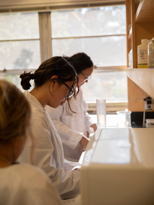 Three young women wearing lab coats stand at a lab bench.