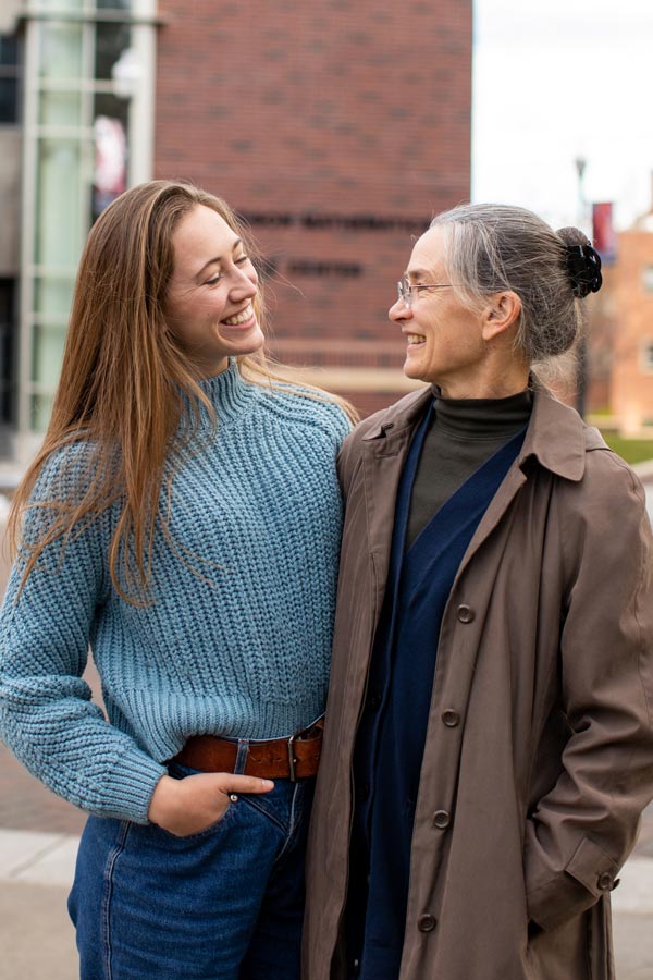 Two women stand side-by-side with their arms around one another and looking at each other. They are both smiling.