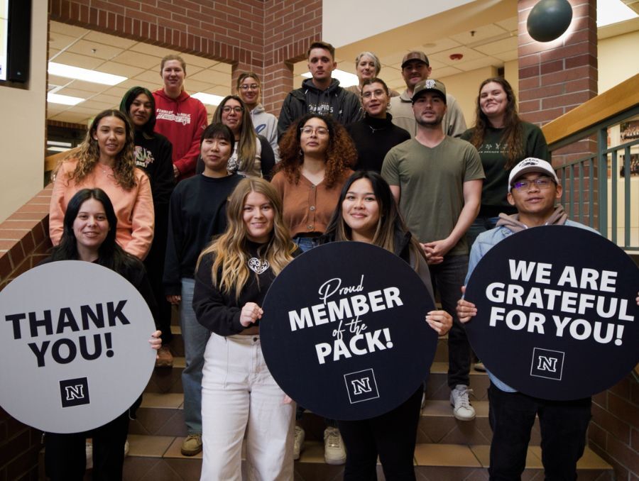 A group of 17 people, mostly students, stand on the steps inside the Reynold's School of Journalism smiling. The front students hold signs saying "Thank you!" and "Proud Member of the Pack!" and "We are grateful for you!"