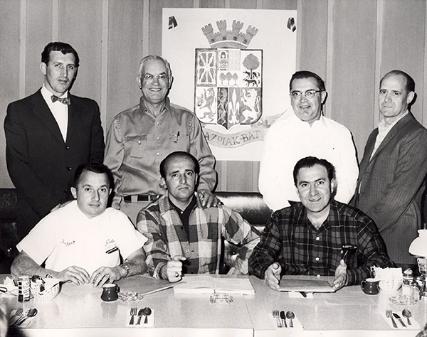 Three men seated at a table with four men standing behind them. Behind the men standing there is a Basque crest hanging in the wall. 
