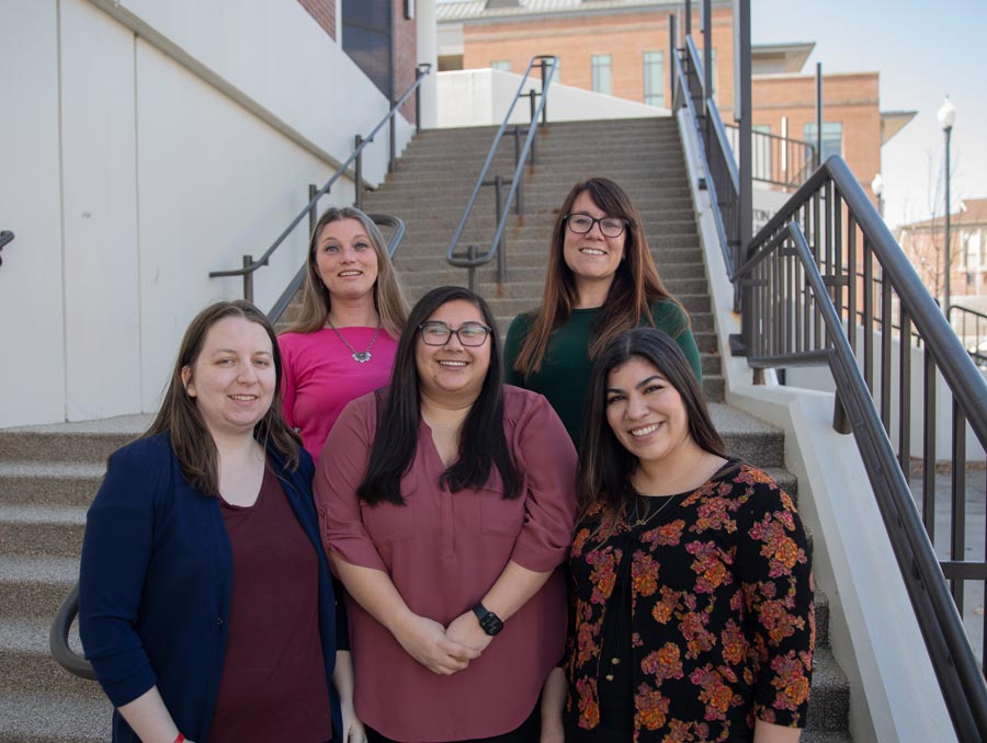 Five women stand at the base of a flight of stairs. Three women stand in the front, two in the back.