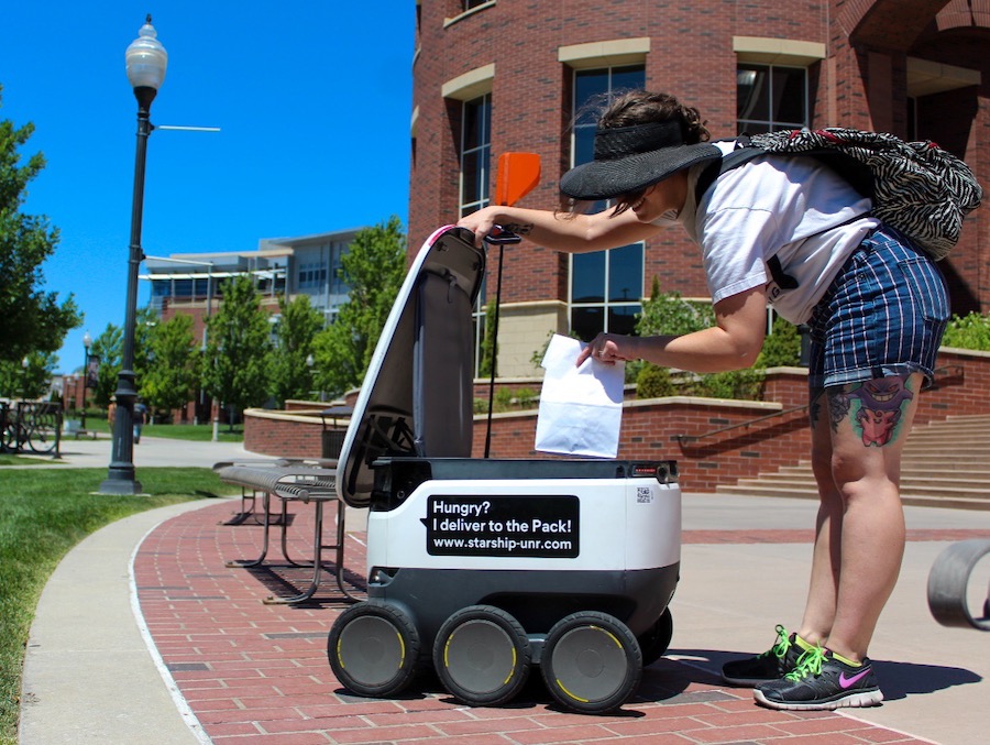On a sunny day outside of the Knowledge Center a person bends down taking a white bag out of a Starship delivery robot.