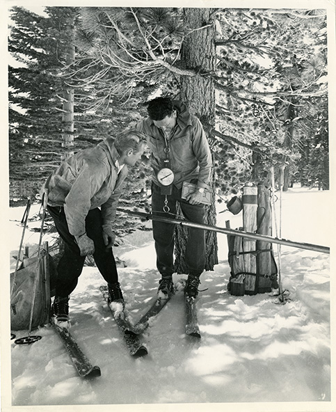 Two men stand in a snowfield wearing skis and winter gear as they take a snow sample to determine how much water content is found in the existing snowpack.