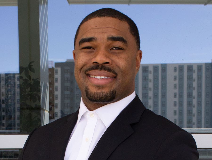 Brandon Sanders smiling in a black suit and white collared button up in front of a glass window that opens up to a blue sky and other concrete buildings. 