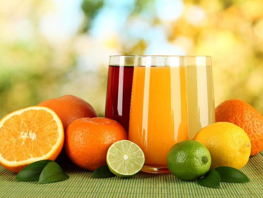 Glass pitchers of colorful fruit juices are surrounded by whole and sliced fruits, such as oranges and limes.