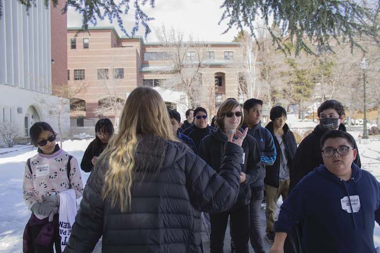 Group of students following a guide with blonde hair who has her back facing the camera, with a brown brick building in the background.