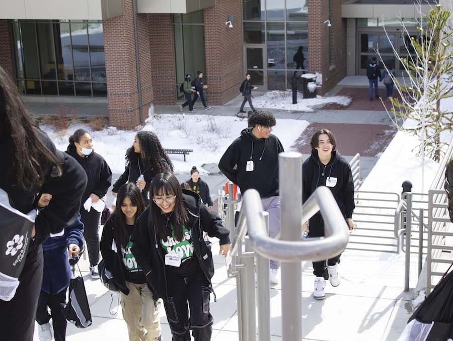 Group of students walking up stairs as they smile and talk amongst each other in front of a brown brick and clear glass building with a silver handrail in the middle.