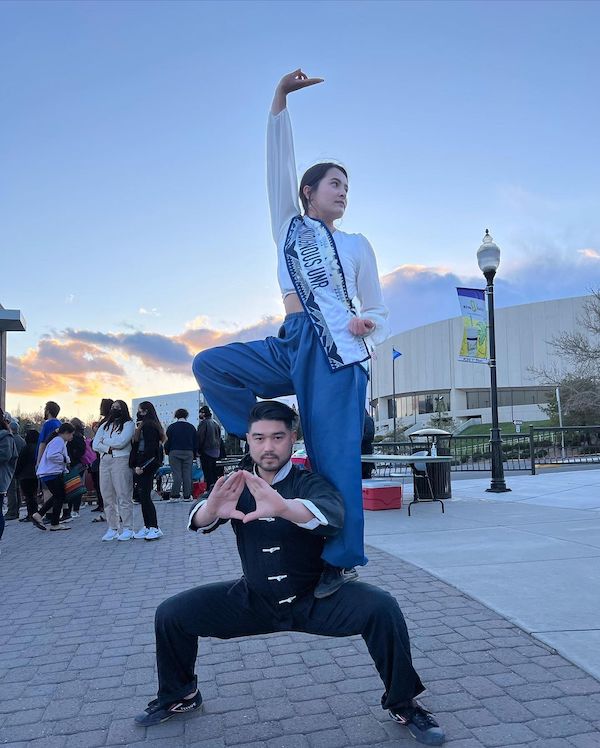 Two performers pose for a photo. The man crouches and a woman stands, posing on top of his leg in a stunt outside in front of the Knowledge Center. 