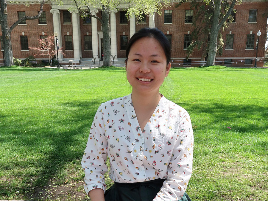 Young woman with dark hair in a white patterned shirt sitting in front of the University quad