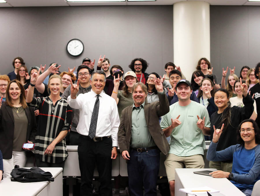 A full classroom of smiling students with President Sandoval standing next to professor Chris Church in the front row.