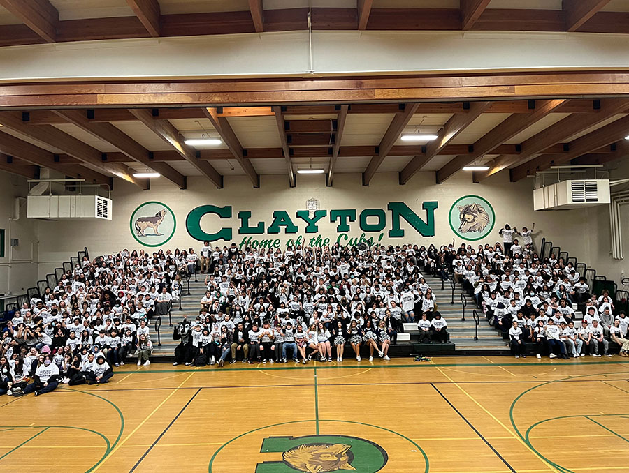 Students sit in bleachers in a gym with the words Clayton painted on the wall behind them.