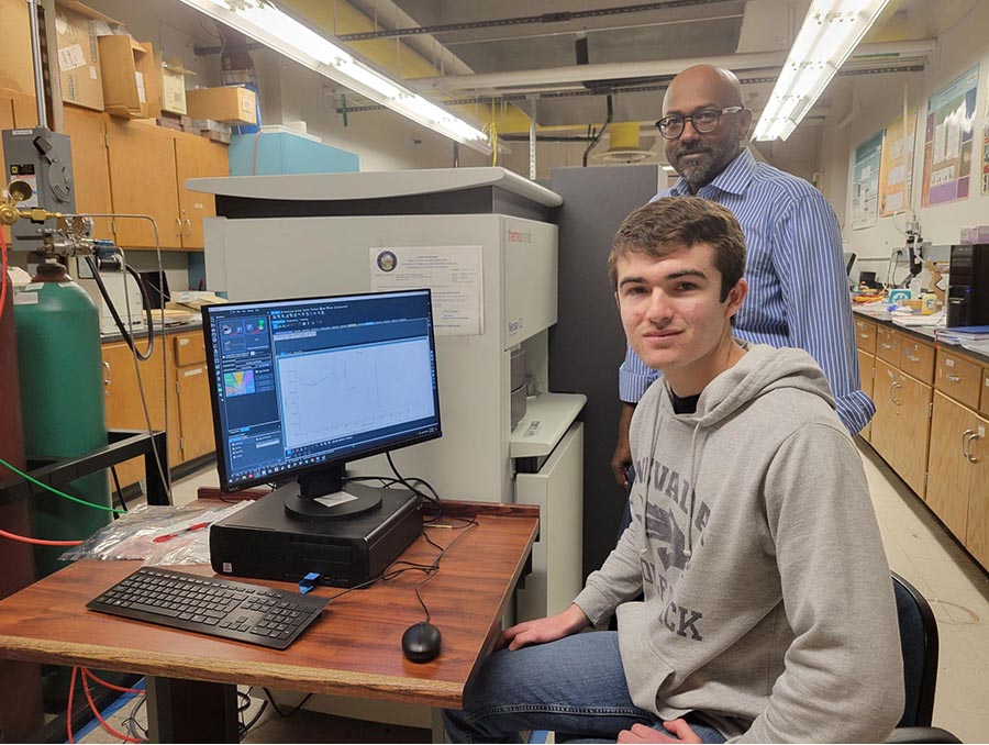 Two people in a lab. A younger man is seating in the foreground at a desk with a computer, looking at the camera. An older man is standing behind him, also looking at the camera.