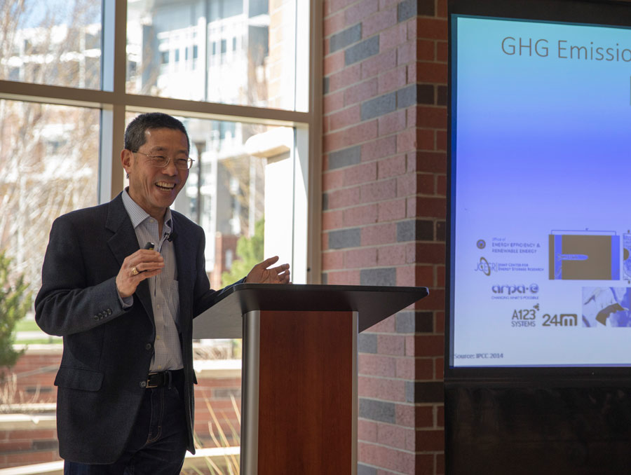 Yet-Ming Chiang stands at a podium laughing. There is a window looking toward the Joe Crowley Student Union behind him, and a projected slide presentation next to him.