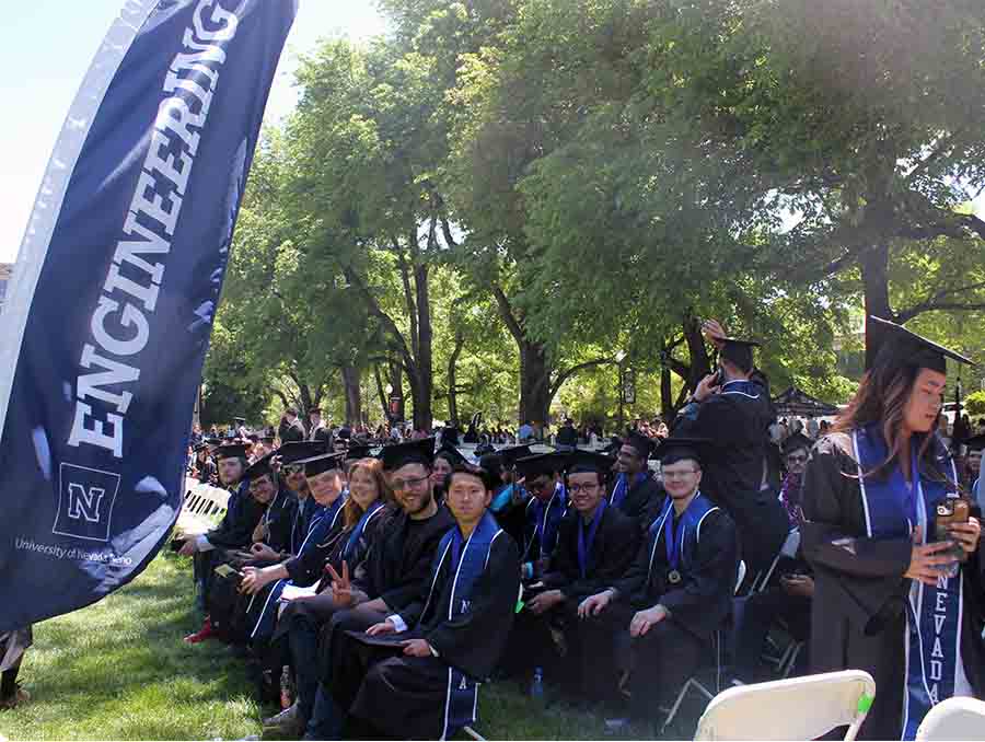 About two dozen graduates in caps and gowns sitting on chairs outdoors on the quad next to a blue banner that reads "Engineering."