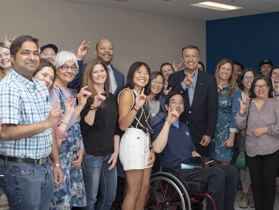 Professor Eric Wang surrounded by a large group of faculty and staff and President Brian Sandoval as they all smile and look at the camera holding up Wolf Pack hand signals.