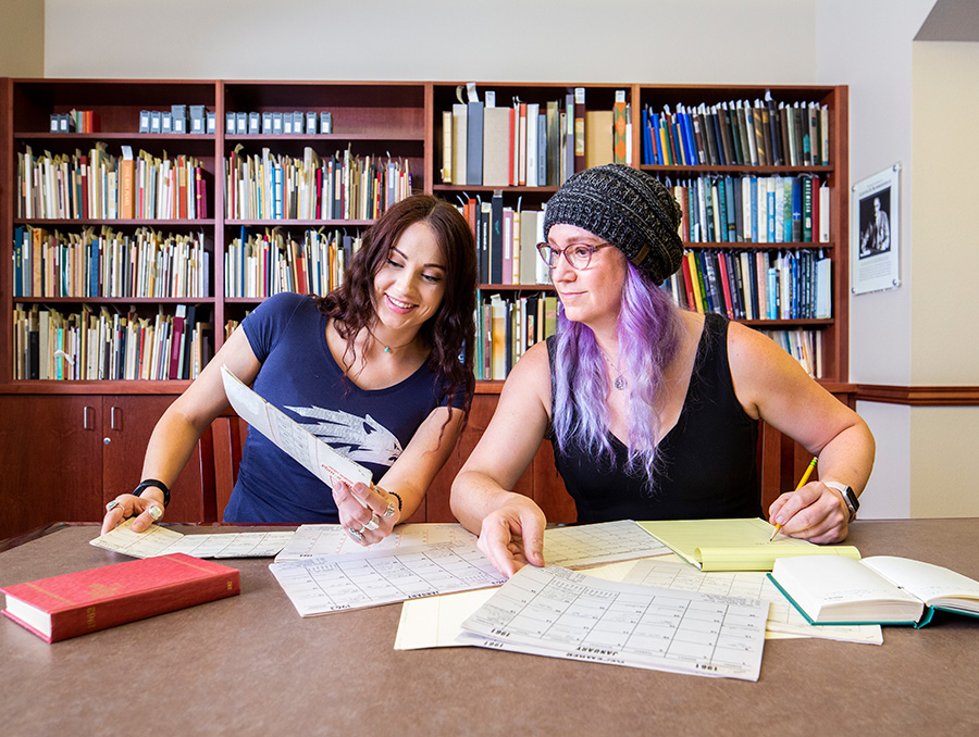 Two women sit at a table inside the Special Collections and University Archives Department. There are folders on the table with historical documents scattered across it.