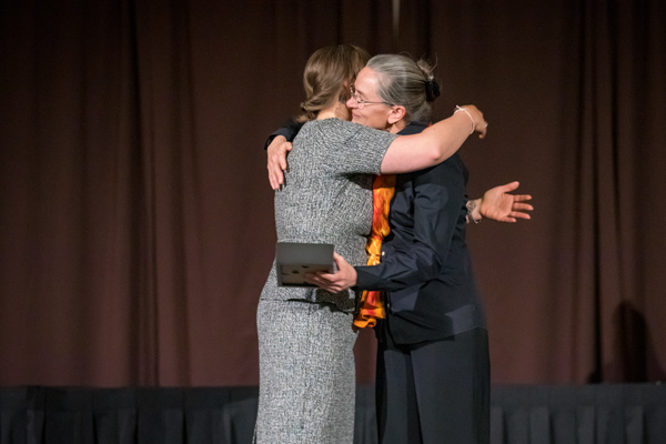 Two women hug on a stage. They are both nicely dressed, and one is holding a framed award.