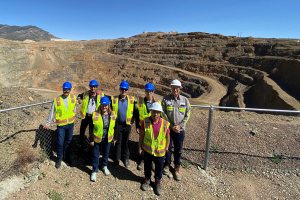 Seven people stand in front of an open pit mine with a fence between them and the edge of the mine.