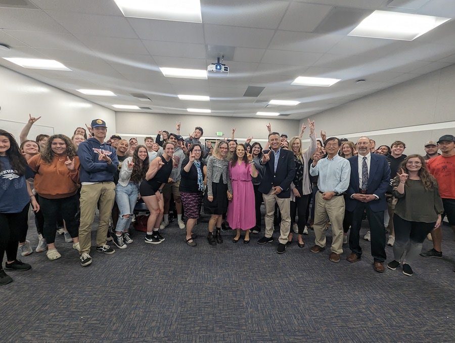 Nora Constantino and President Sandoval pose with a large classroom full of smiling people as she accepts her award announcement.