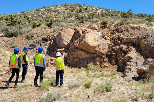 Four people wearing hard hats and high-visibility vests stand in a desert landscape, as one person points at rocks in front of them.