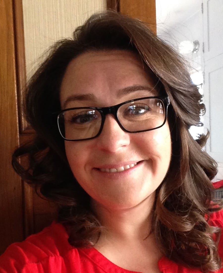 Woman with brown, curled hair stands in front of a brown and glass door. She is wearing a bright red blouse and black, square glasses.