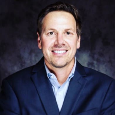 Man in front of a black and white marble backdrop is dressed in a dark, navy blue suit jacket with a light, powder blue button-up shirt underneath.