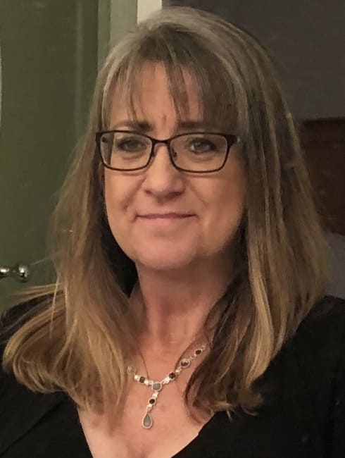 Woman wearing a lower cut black blouse with a necklace. She has dirty blonde hair and bangs. 