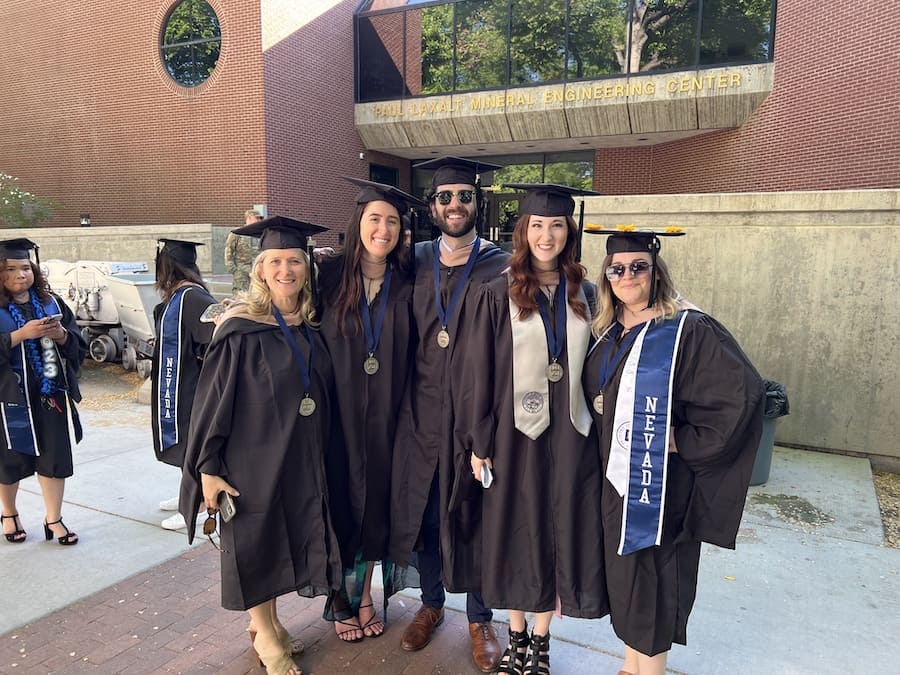 A group of 7 master's students wearing graduation regalia stand smiling outside, posing for the camera.