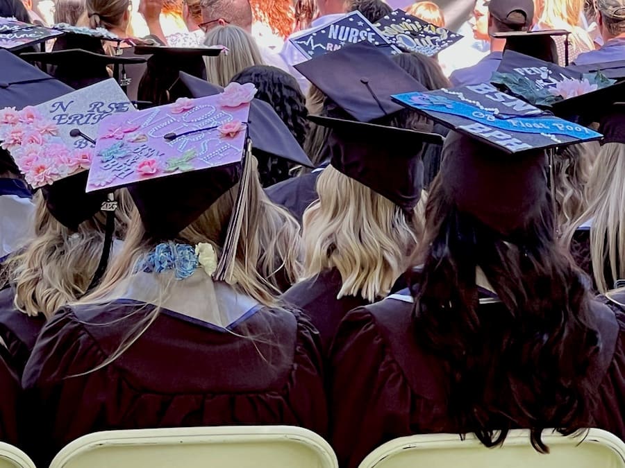 The backs of various grads are seen, some with decorated graduation caps for Nursing, as they look to the commencement stage.