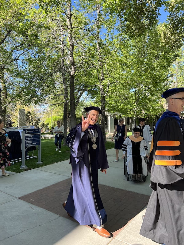 President Sandoval walks by in graduation regalia as he makes the Wolf Pack hand signal, smiling.