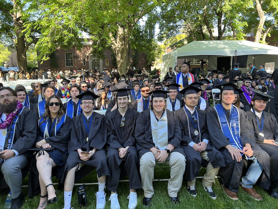 A large group of grads sit before the commencement ceremony starts wearing their graduation caps and gowns and smiling broadly in the sunshine.