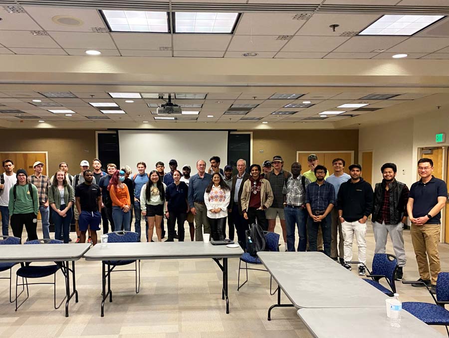 A group of around 30 people stand in a classroom with tables in front of them.