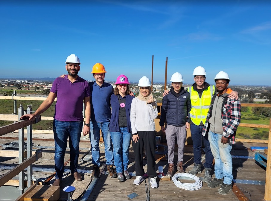 A group of researchers stands on a sunny roof wearing hardhats and smiling at the camera.