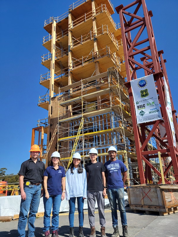 A group of four researchers stand at the bottom of the TallWood building smiling and wearing hardhats.