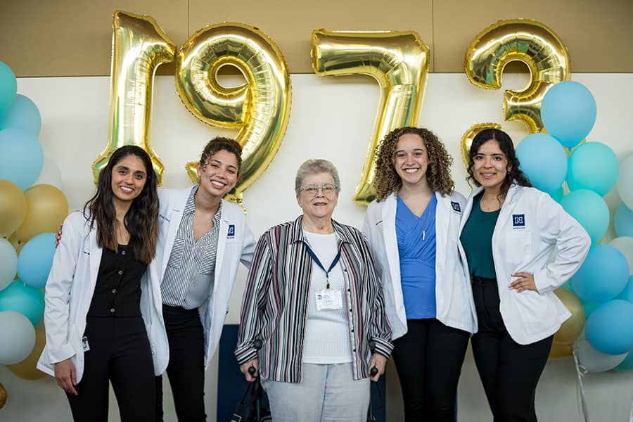 Five women, most in white UNR Med lab coats, smile and pose for the camera in front of the 1973 balloon display for the time capsule reveal
