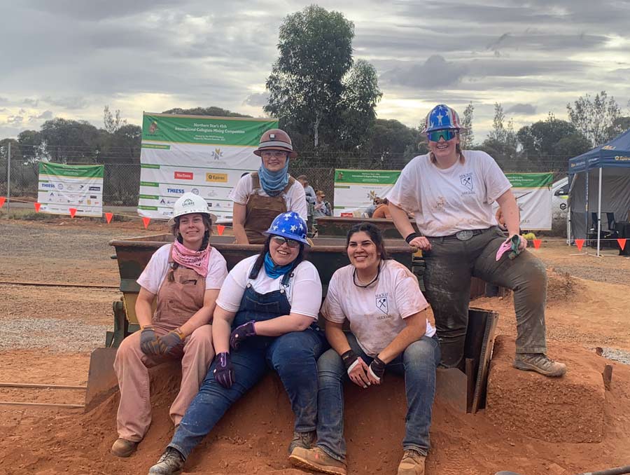The Mackay Muckers Women's Team sits on a pile of dirt in front of a mining cart. They are all covered in dirt.