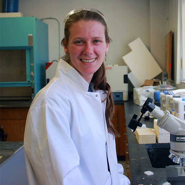 Abbey Hageman sits at a desk with a microscope, looking at the camera