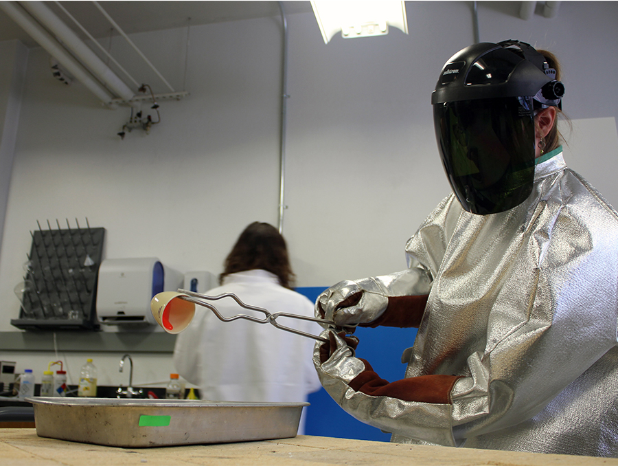 Abbey Hageman in a lab wearing lab coat, gloves and face protection, using tongs to hold a small cup with pink liquid, which she is pouring into a pan. Another person, facing away from the camera, is in the background.