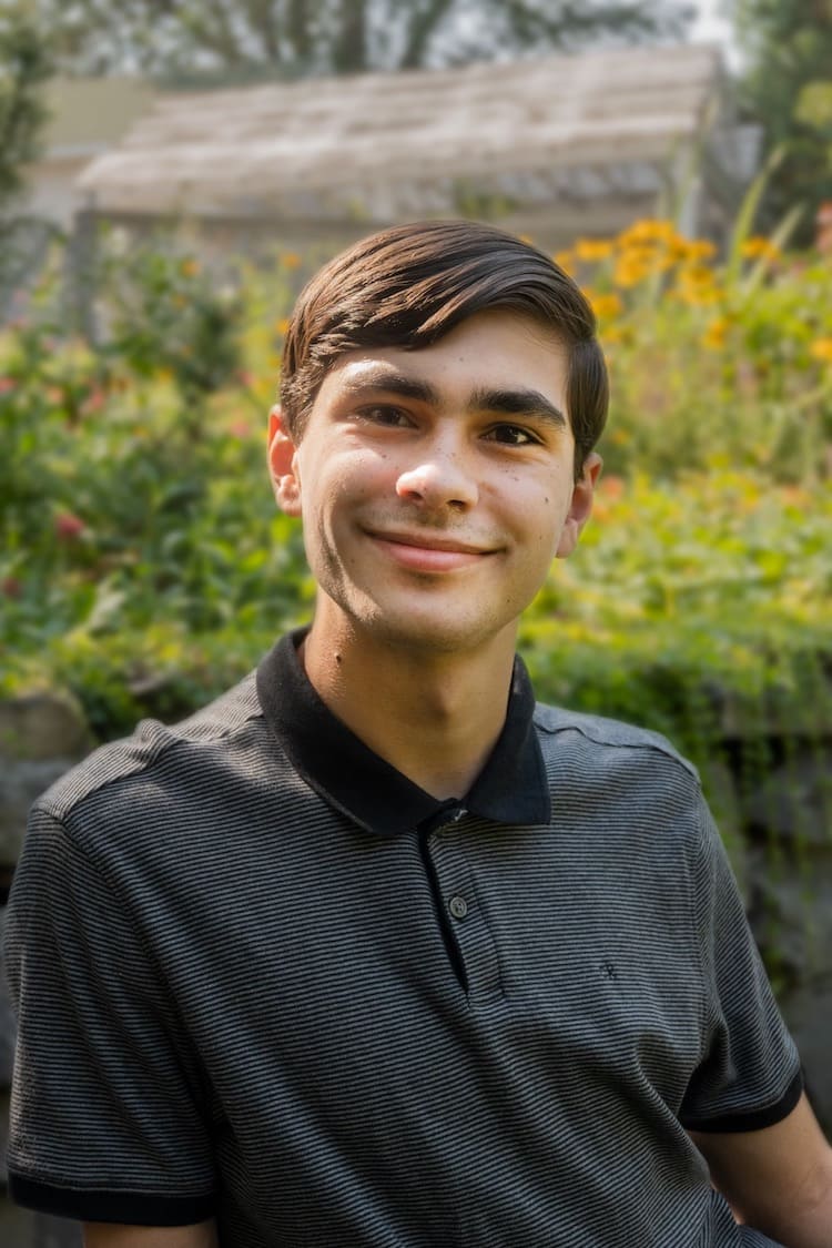 Man smiling and wearing a gray collared shirt sitting in front of a large bed of flowers.