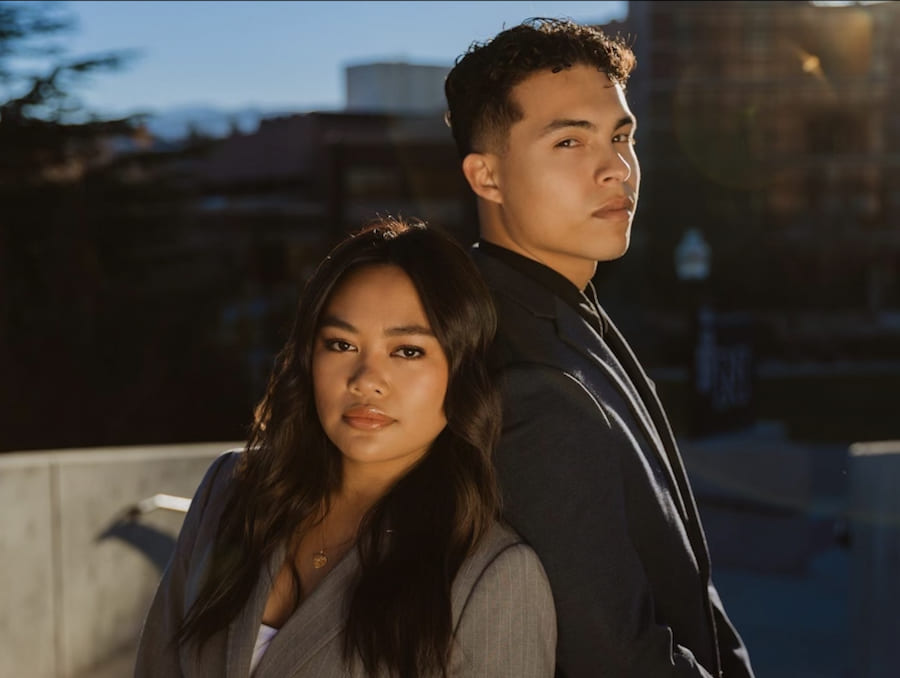 Woman in a beige suit jacket and white shirt stands back-to-back with man in a black suit jacket and black button-up shirt. The two stand in front of a gray wall with trees and a tall building in the background.