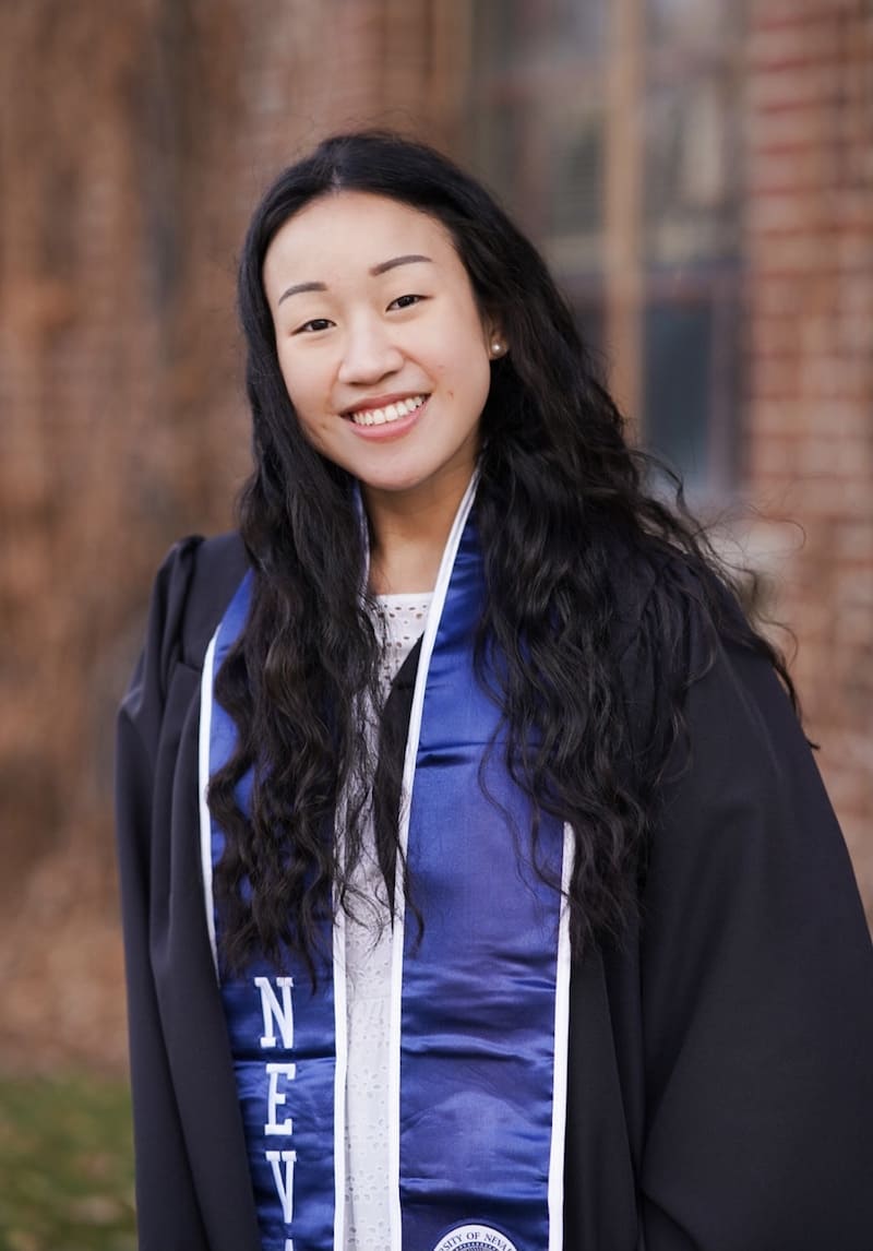 Woman in white dress with black graduation gown and blue graduation stole. She is standing in front of a blurred out brown building. 