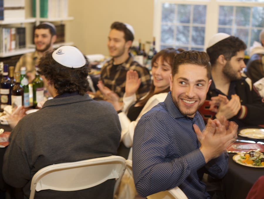 Group of people sitting around a dinner table. Some of the men are wearing white Yamakas on their heads. 