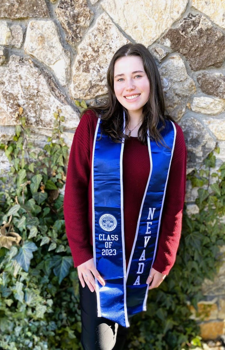 Woman in a red long sleeve shirt and black pants holding a blue graduation stole that says, "Class of 2023" on one side and "NEVADA" in a vertical lettering on the other side.