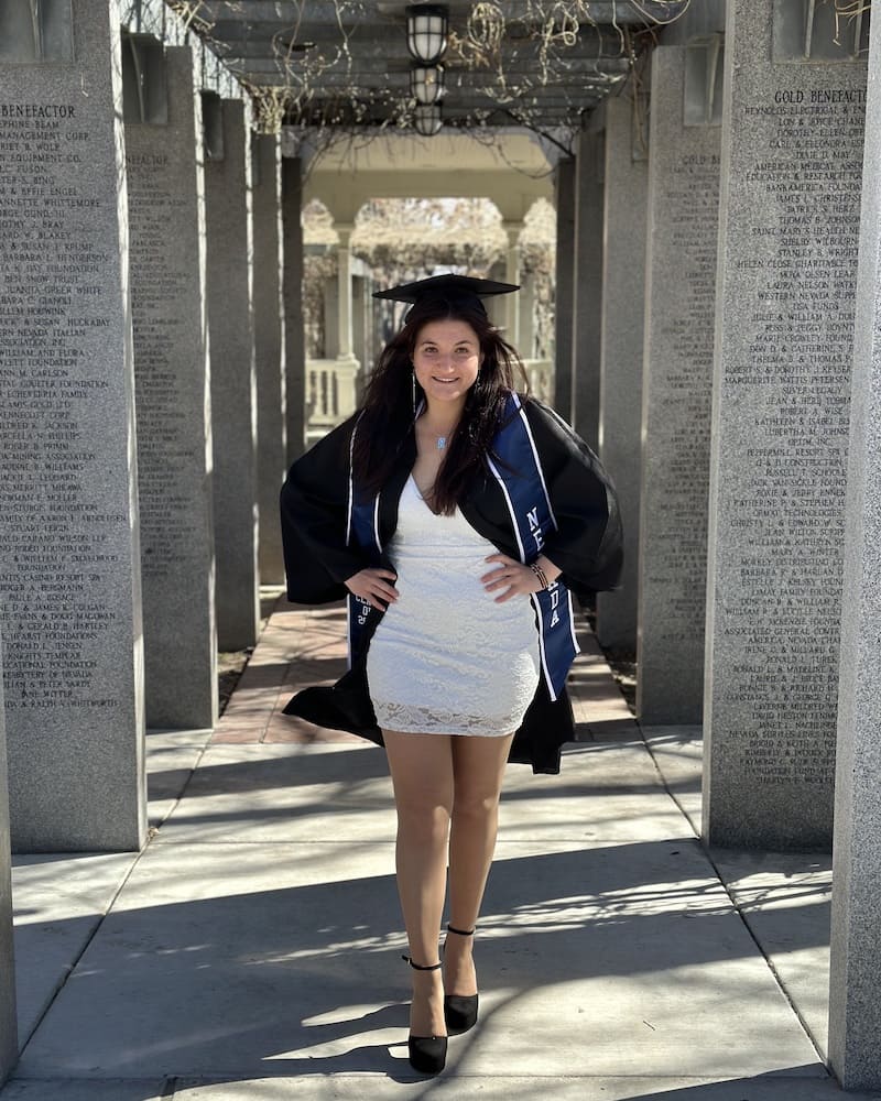 Woman in a white dress and black graduation cap and gown posing with her hands on her hips. She is standing in between gray, concrete pillars that have writing etched into them.