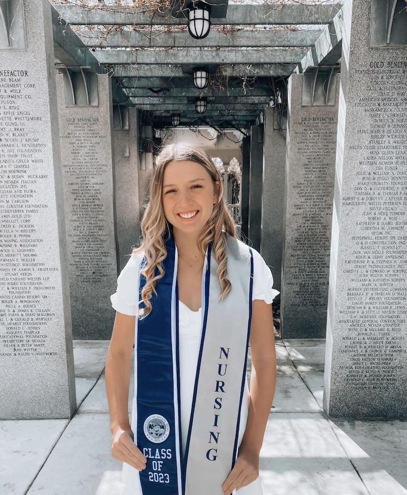 Woman in a white dress covered in a blue and silver graduation stole. One side says, "Class of 2023" and the other side says "NURSING" in vertical lettering. She is standing in the middle of gray pillars that have names etched into them.